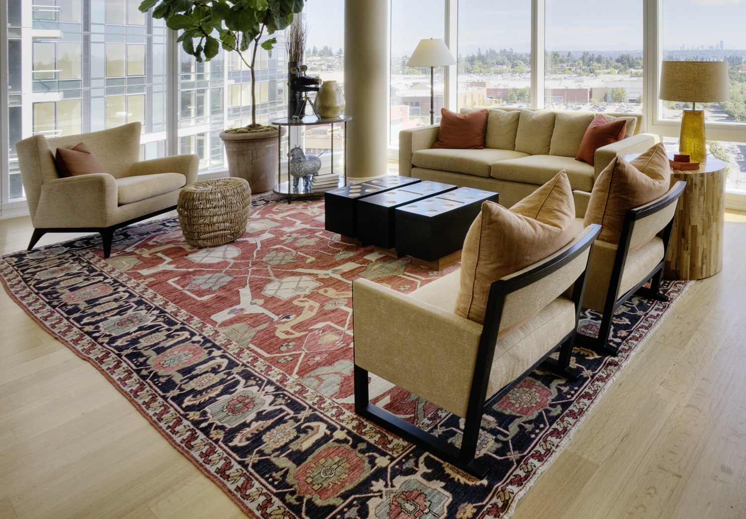 A modern living room with large windows, beige furniture, a red patterned rug, a potted plant, and a cityscape view in the background.