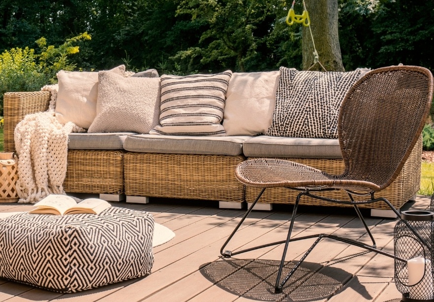 Outdoor seating area with a wicker sofa, multiple cushions, a patterned pouf, a brown metal chair, a blanket, and an open book on a wooden deck surrounded by greenery.