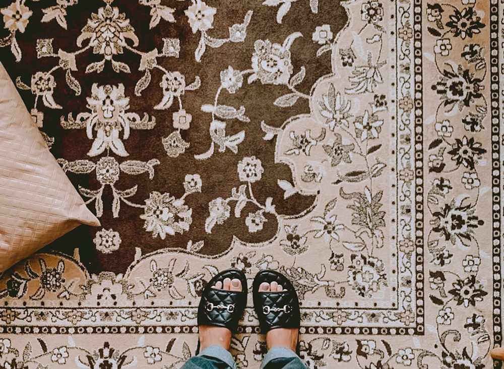Person wearing black sandals stands on an ornate, patterned rug with floral designs; a beige cushion is partially visible on the left.