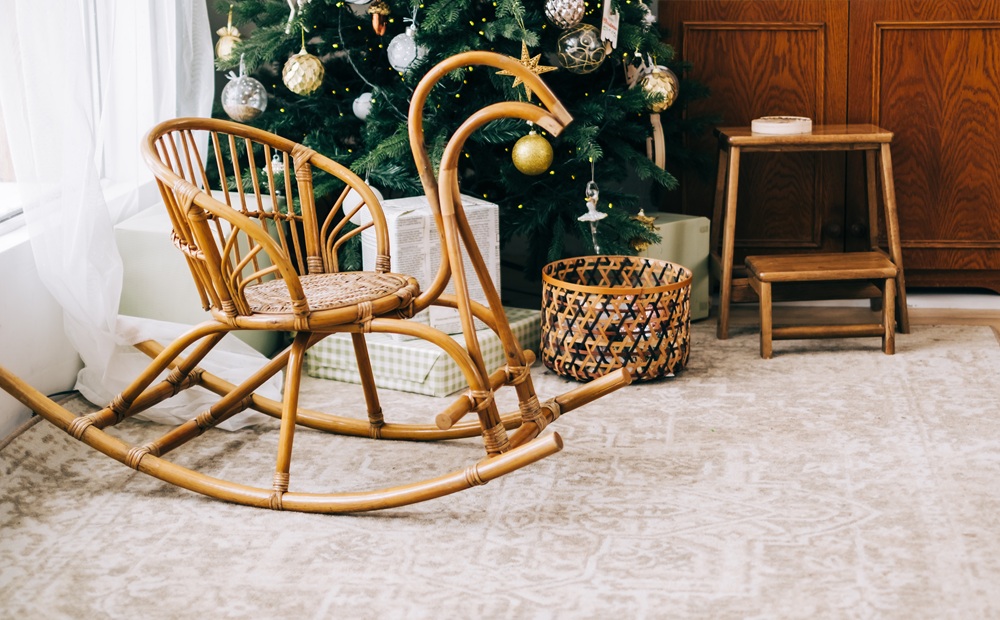 A wooden rocking chair sits on a patterned rug near a Christmas tree decorated with ornaments, with a basket and small wooden table nearby.
