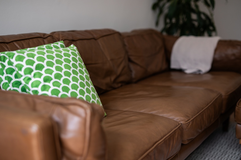 A brown leather couch with a green patterned pillow and a light-colored blanket draped over the armrest.