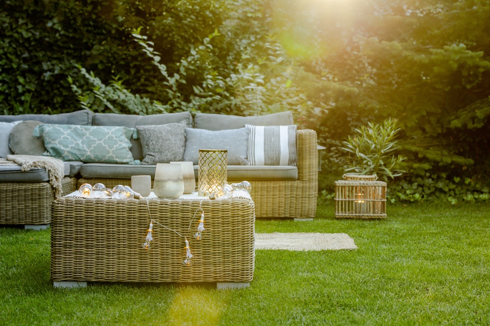 A wicker outdoor sofa and table with cushions and candles are set on a grassy lawn, surrounded by greenery, in warm sunlight.