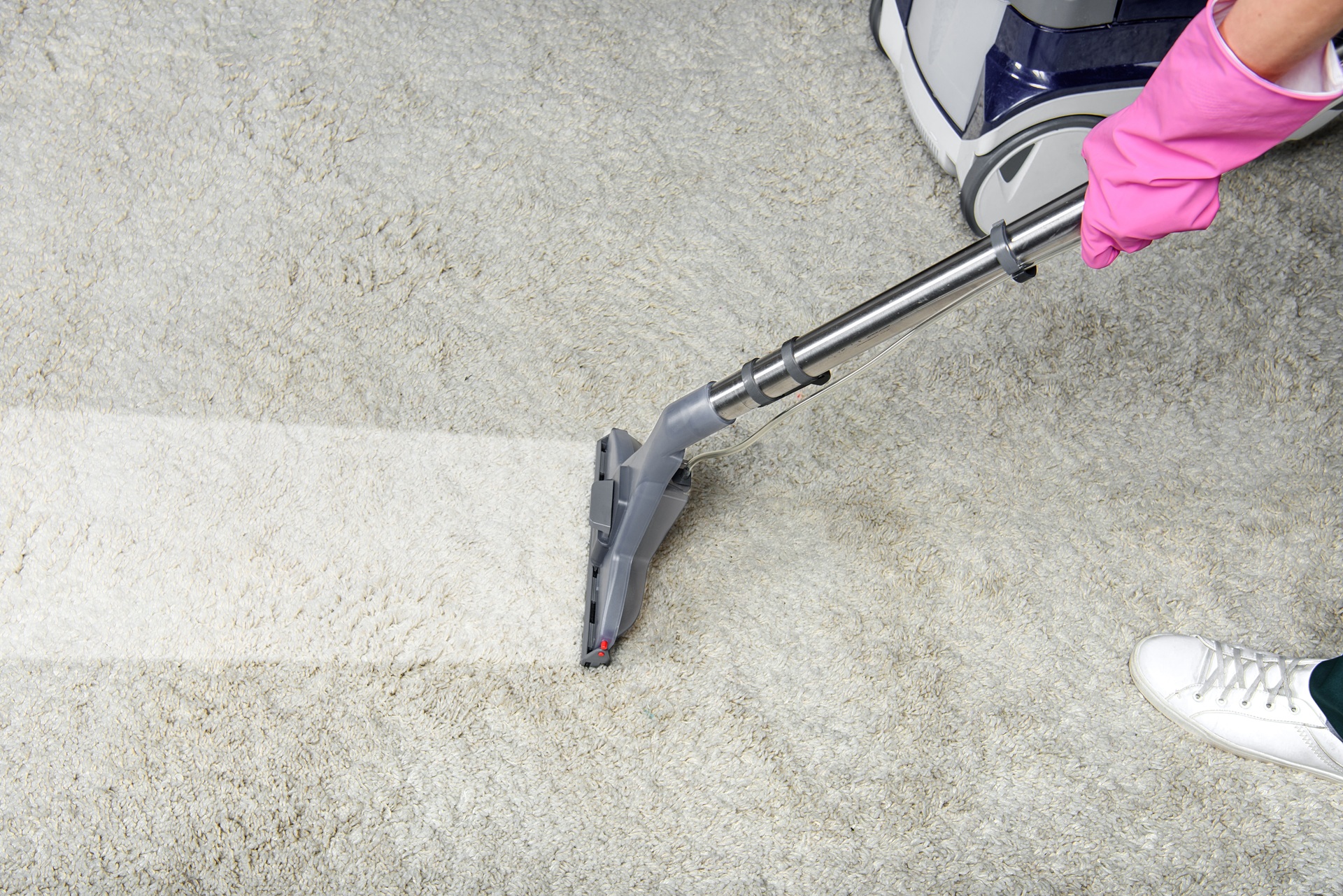 A person wearing a pink glove uses a carpet cleaning machine on a light-colored carpet, showing a visibly cleaner path.