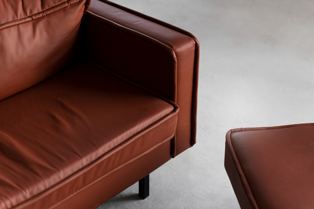 Close-up view of a brown leather armchair with visible stitching, positioned next to a matching leather ottoman on a light-colored floor.